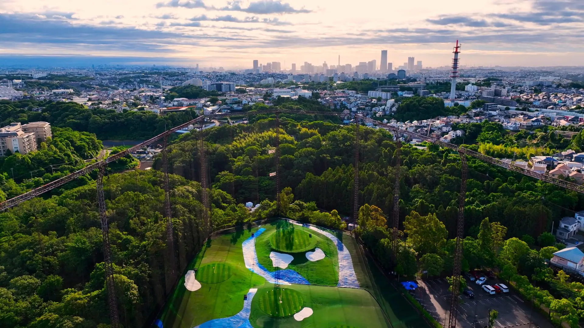 夕暮れのHANZ GOLF CLUBを上空から見た全景。谷に沿って芝の外野が広がり、両側に打球ネットの支柱が立ち、奥に横浜みなとみらいの街並みが見える。